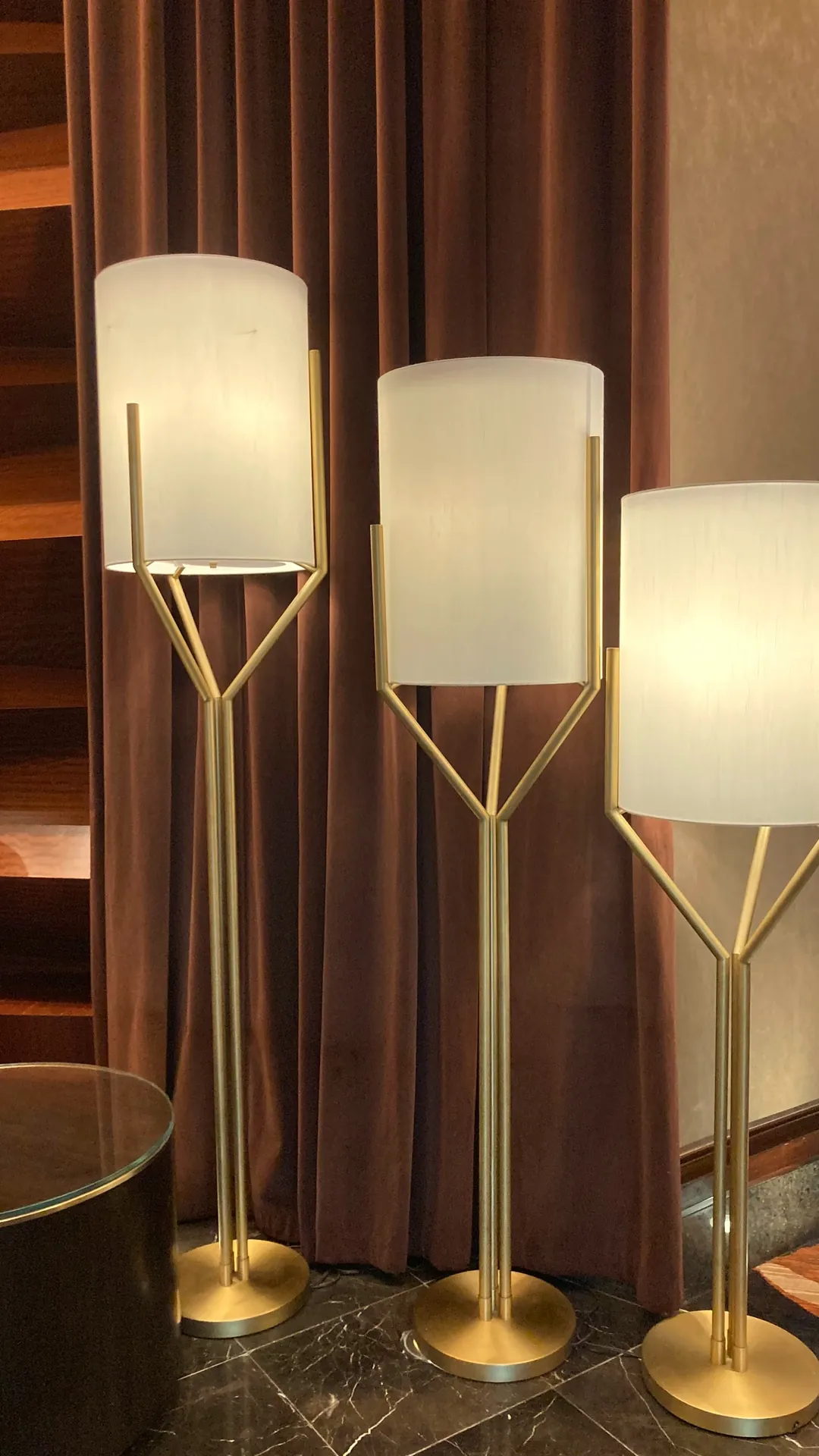 Three brass floor lamps with white shades in descending heights in front of burgundy drapes