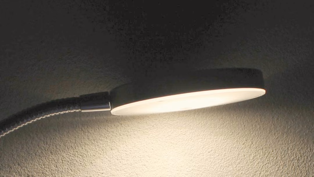 Slim LED disc lamp on a gooseneck arm, lit, casting light across a textured wall