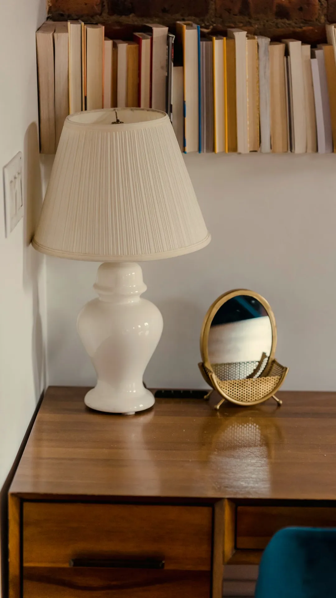 White table lamp with a pleated shade on a small wooden desk below a shelf of books