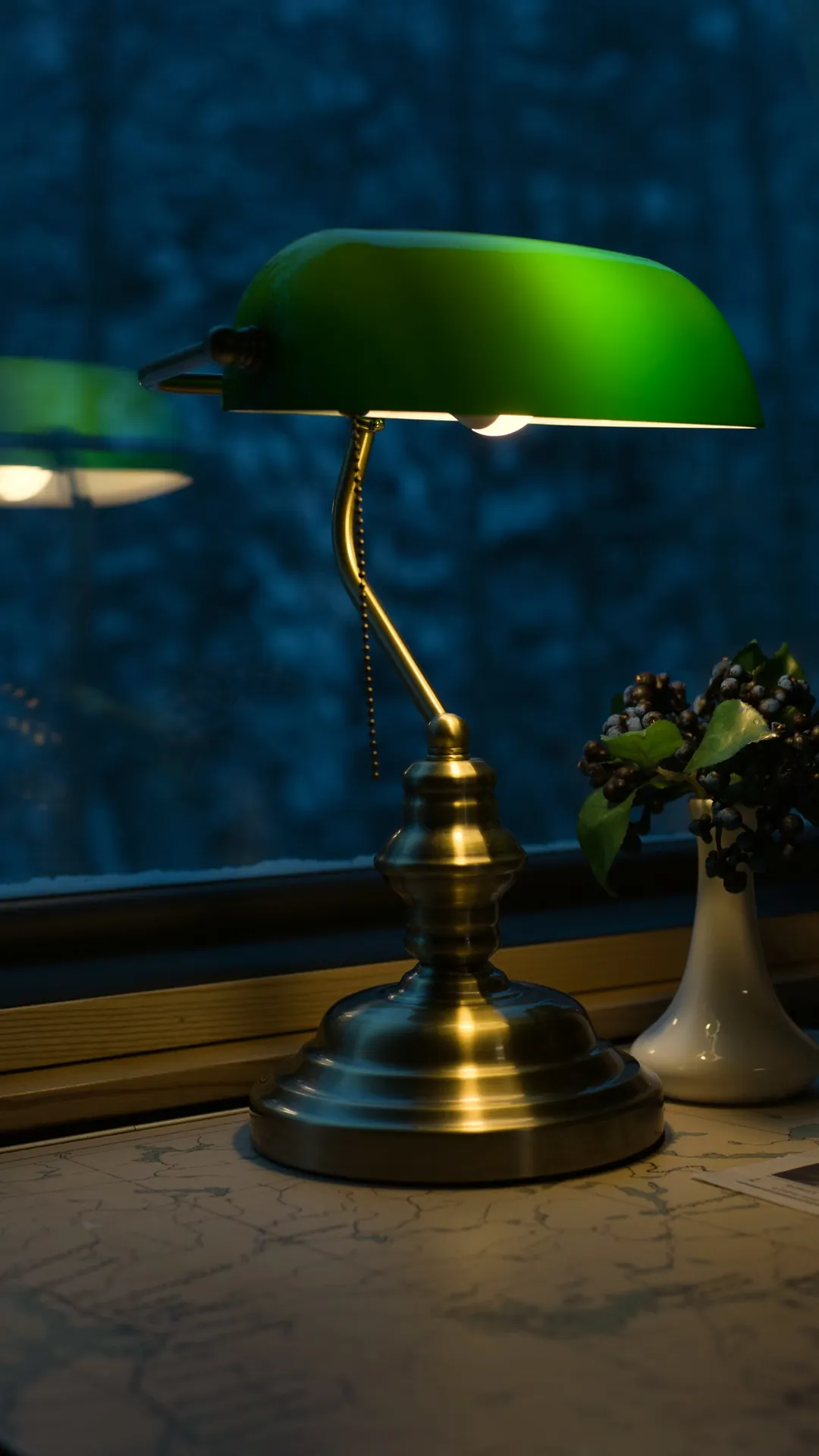 Brass banker lamp with a green glass shade on a desk by a window at dusk