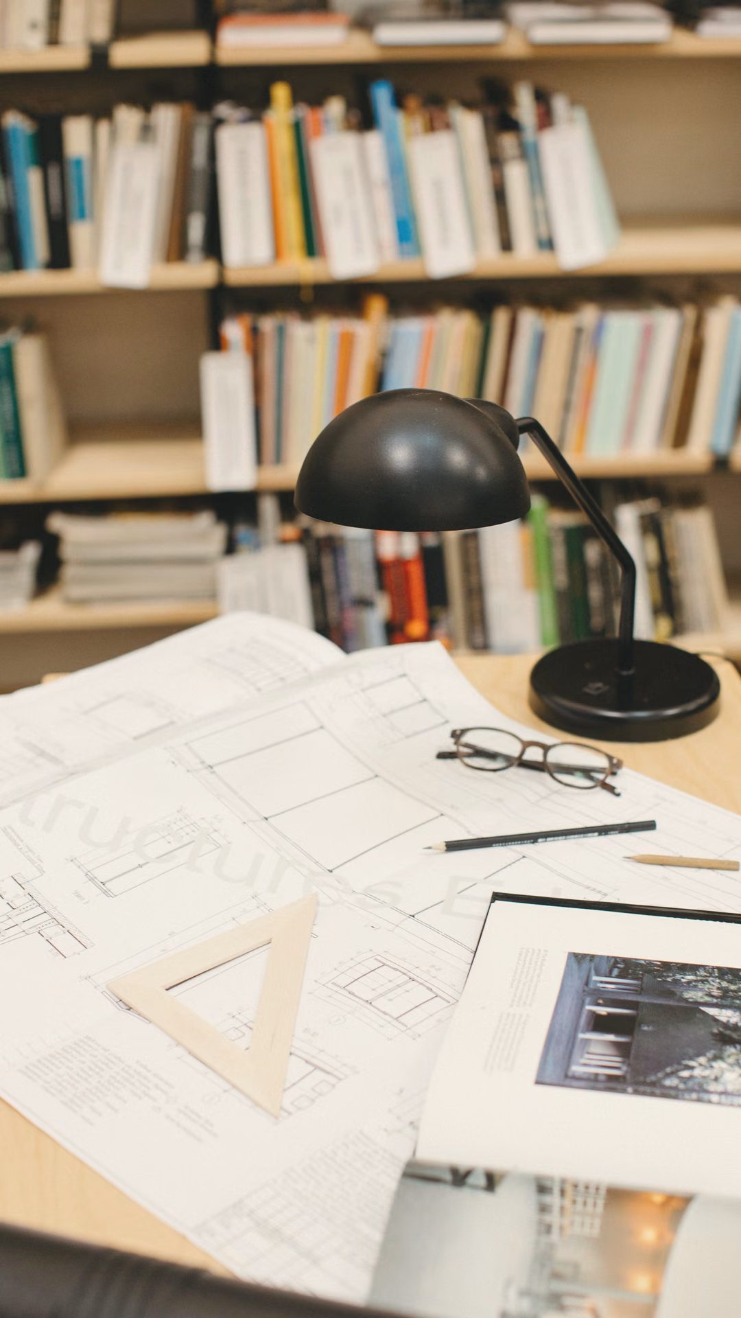Black desk lamp lighting drawings on a drafting desk in front of bookshelves