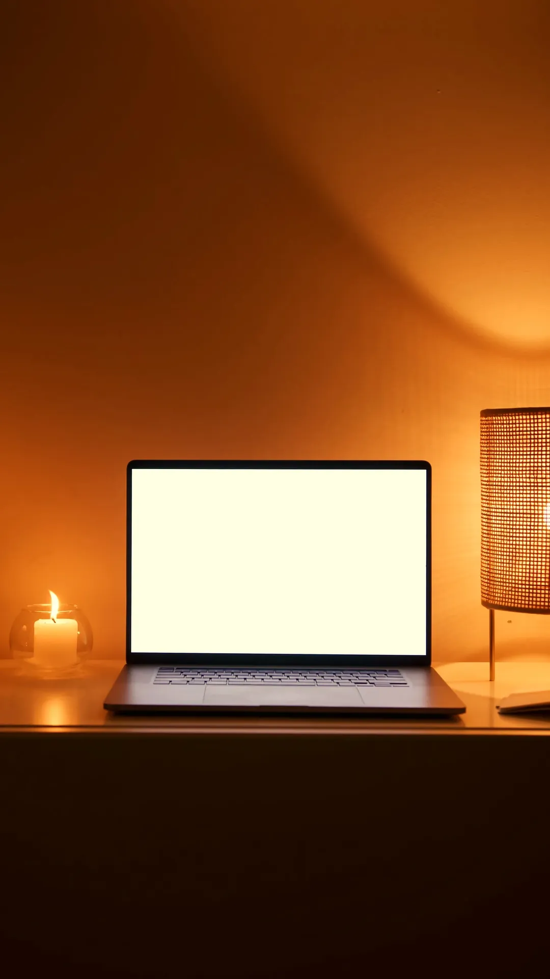Laptop with a blank bright screen on a desk beside a woven table lamp and a candle in warm light